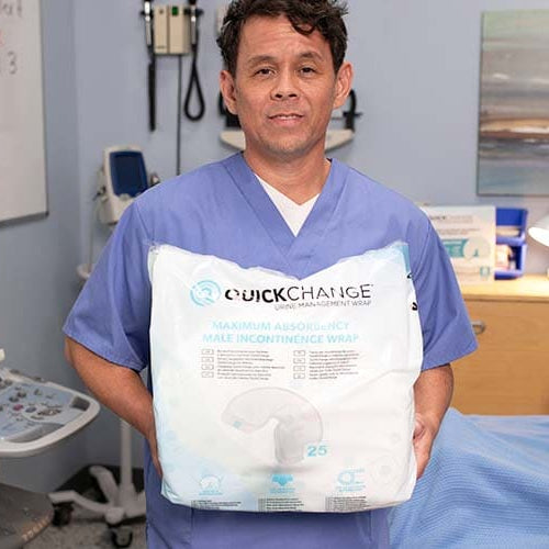 A man in blue medical scrubs holds a large package of QuickChange Maximum Absorbency Wipe incontinence wraps in a clinical room setting.