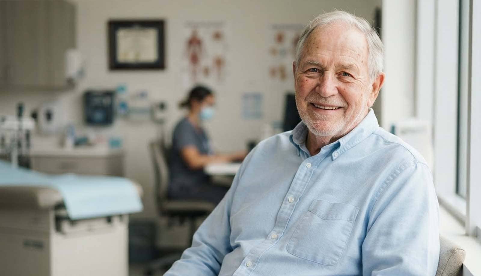 A smiling older man in a light blue shirt sits in a bright clinic room.