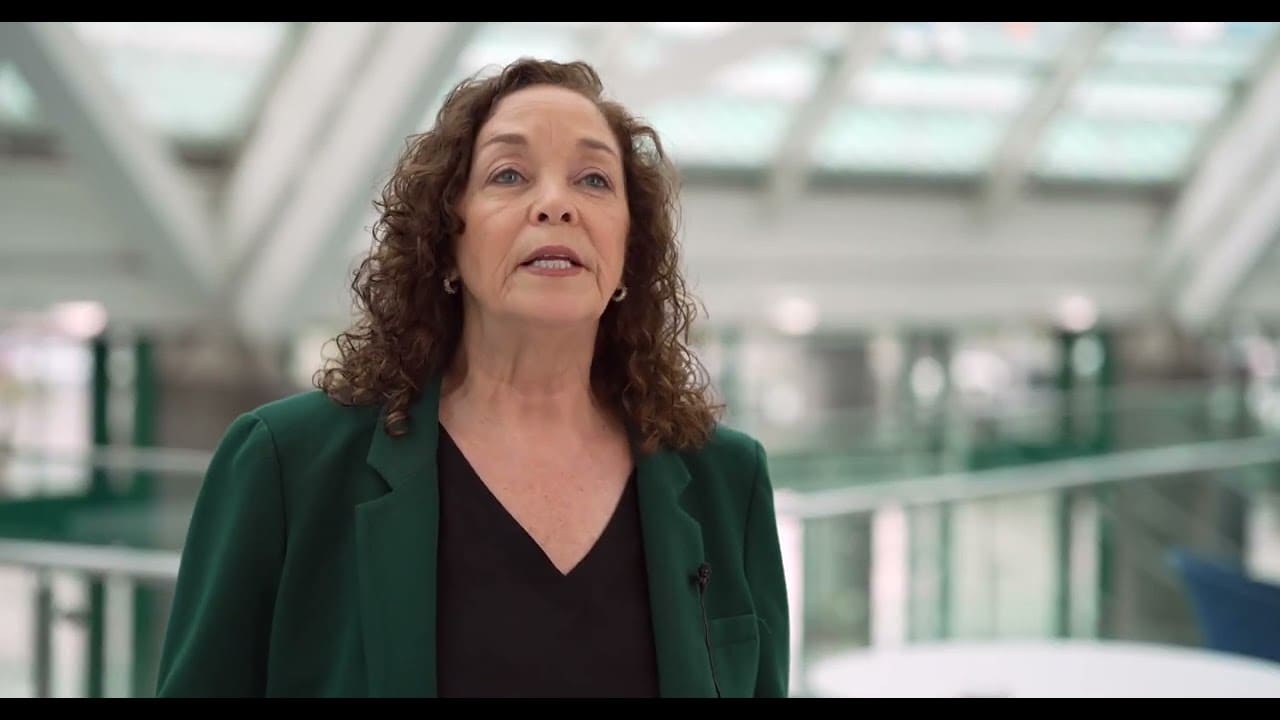 A woman with curly brown hair wearing a dark green blazer over a black top, standing in a modern indoor setting with a blurred glass railing and ceiling behind her.