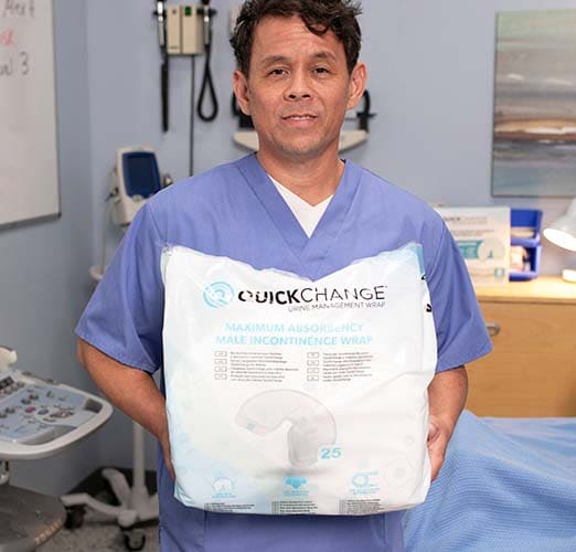 A man in blue medical scrubs holds a large package of QuickChange Maximum Absorbency Wipe incontinence wraps in a clinical room setting.